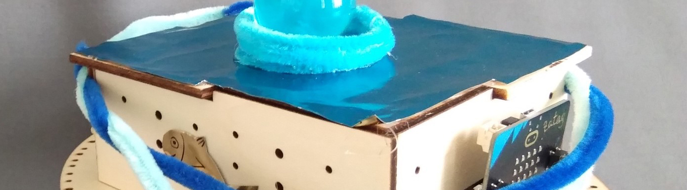 "Unterwasser-Tierärztin-Roboter" aus einer runden Holzplatte mit Rädern, auf der Platinen und ein eckiger Holzaufbau sitzt. Dekoriert ist der Roboter mit hell- und dunkelblauen Pfeifenputzern und hat eine blaue Glitzeroberfläche. Eine hellblaue glitzernde Glasmurmel sitzt mittig auf der Deckplatte.