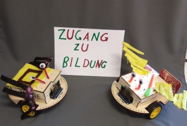 Von Schüler*innen gebaute Bildungsroboter. Die Roboter stehen links und rechts neben einem Schild mit der Aufschrift "Zugang zu Bildung". Beide Roboter haben eine runde Bodenplatte, zwei Räder und sind liebevoll dekoriert. Beide Roboter haben Augen. Der linke Roboter ist in schwarz-gelb dekoriert und der rechte Roboter hat eine Karotte auf der Deckenplatte und gelbe Streifen.