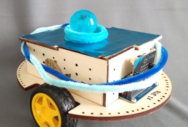 "Unterwasser-Tierärztin-Roboter" aus einer runden Holzplatte mit Rädern, auf der Platinen und ein eckiger Holzaufbau sitzt. Dekoriert ist der Roboter mit hell- und dunkelblauen Pfeifenputzern und hat eine blaue Glitzeroberfläche. Eine hellblaue glitzernde Glasmurmel sitzt mittig auf der Deckplatte.