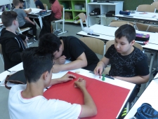Drei Burschen sitzen an einem Tisch und zeichnen ihr Game Design auf ein großes rotes A2-Blatt, "Game Design", ZIMD