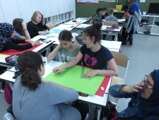 Buben und Mädchen sitzen in Kleingruppen an Tischen und arbeiten an ihren Spielen. Sie haben Buntpapier, Lineal und Stifte auf ihren Tischen. "Game Design", ZIMD