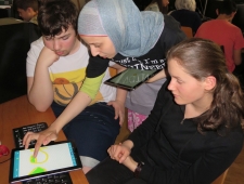 Ein Bursche und zwei Mädchen sitzen um ein Tablet. Das Mädchen in der Mitte zeichnet eine Grafik mit dem Finger auf dam Tablet, "Pocketcode", ZIMD