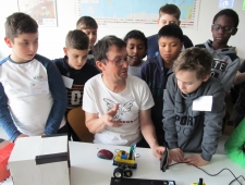 Roberta, ZIMD Andreas Landl sitzt an einem Tisch. Eine Gruppe Buben versammelt sich um ihn. Er erklärt die Verkabelung des Roboters, der vor ihm auf dem Tisch steht. "Roberta", ZIMD