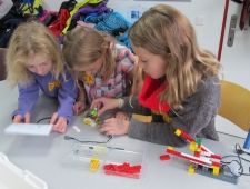 Drei Mädchen arbeiten gemeinsam an ihrem bunten Roboter aus Lego. Das Mädchen links, liest die Bauanleitung dazu. "Robina", ZIMD