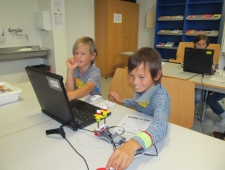 Zwei Buben sitzen vor dem Laptop bei der Programmierung ihres selbst gebauten Roboters, der verkabelt vor ihnen auf dem Tisch steht. Beide Buben lächeln. "Robina", ZIMD